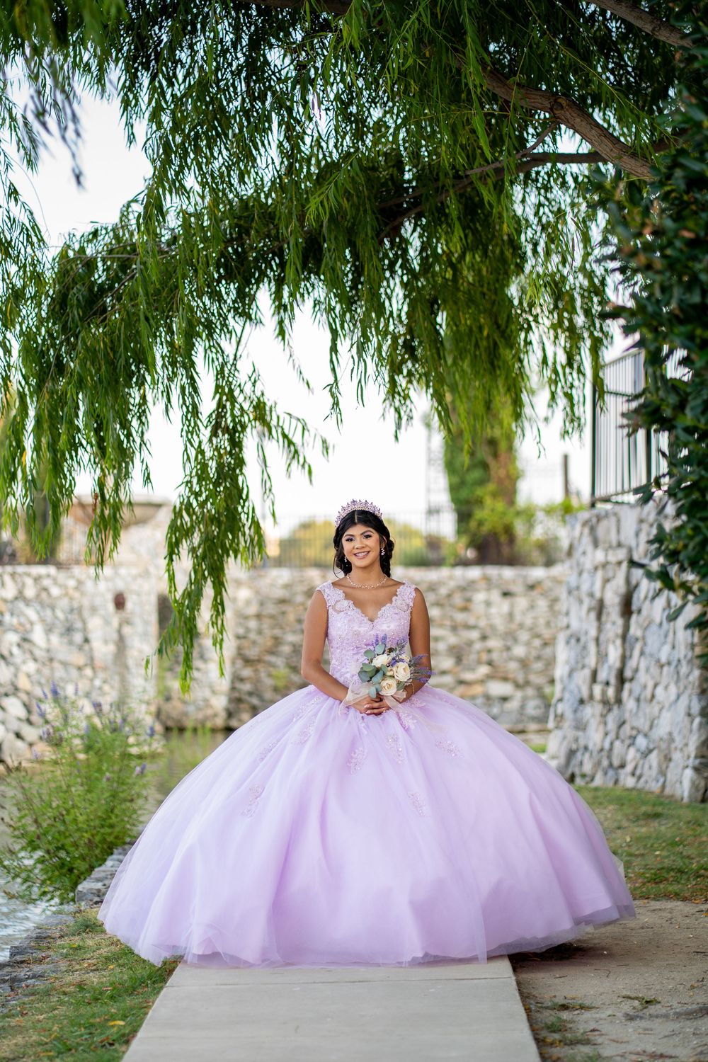 Stunning quinceañera portrait with flowing pink princess ball gown under weeping willow trees.