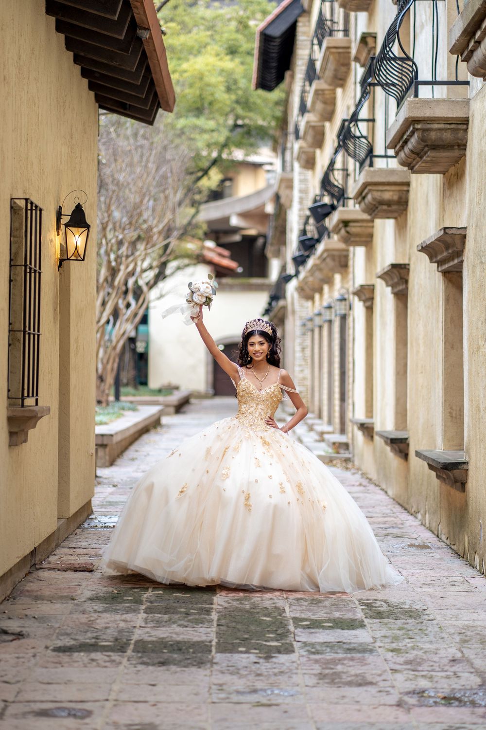 A stunning quinceanera dress flows elegantly along a stone colonnade walkway with ornate lanterns.