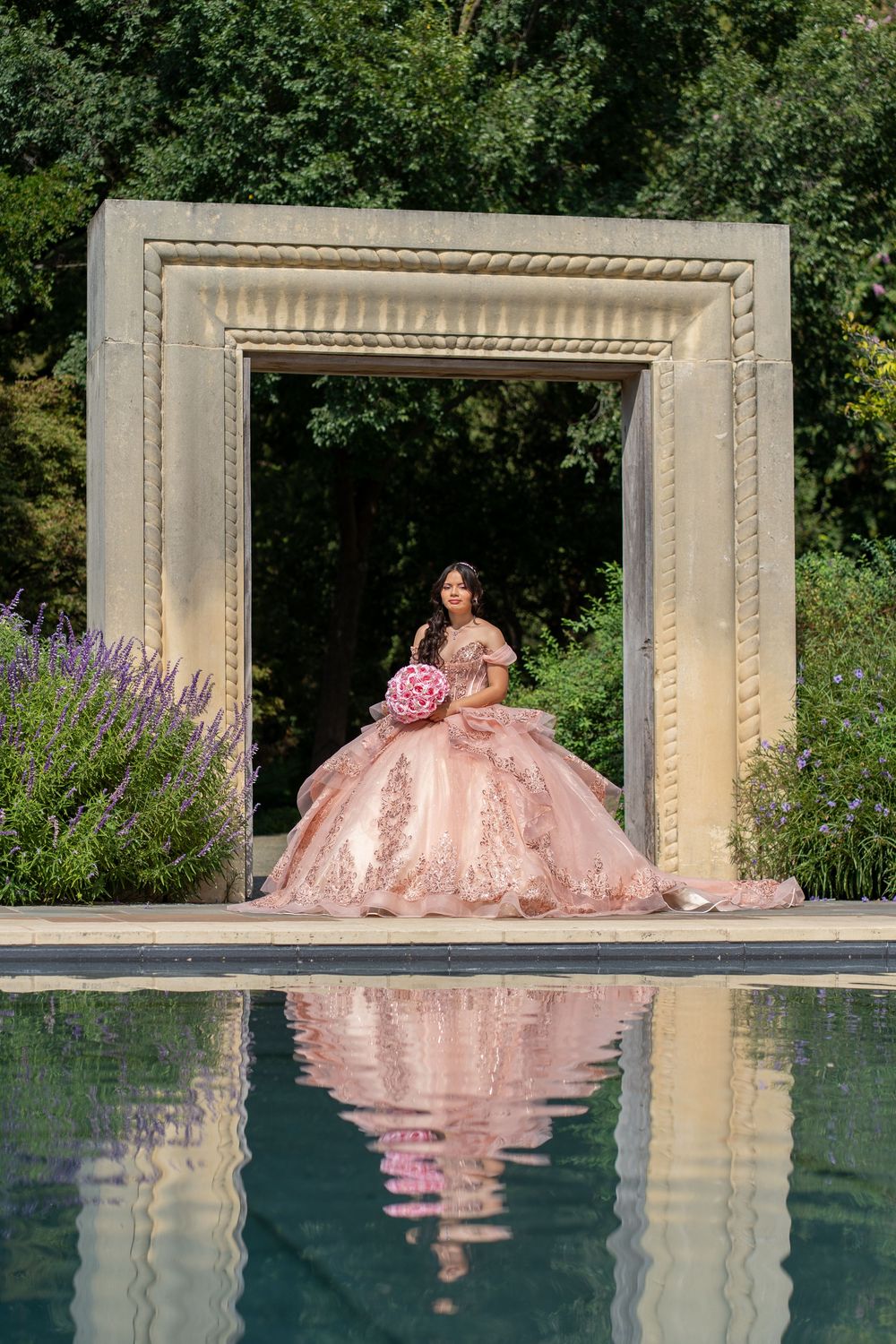 A pink ballgown reflects beautifully in a pool with decorative stone archways and purple flowers.