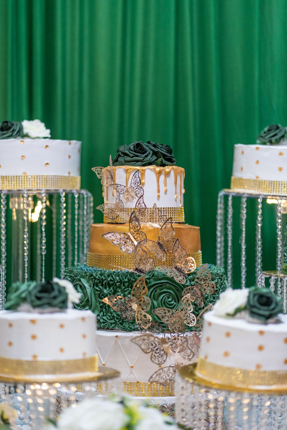 Luxurious white and gold tiered cakes with butterfly decorations against a green backdrop.