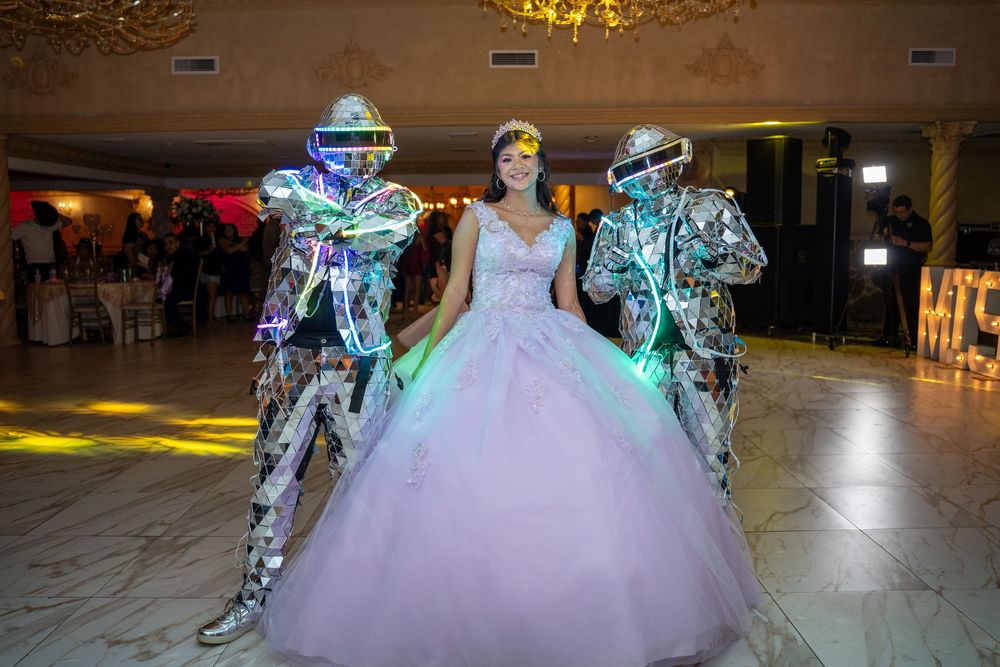A person in a flowing white quinceañera dress spins gracefully on a dance floor under colorful lighting effects.