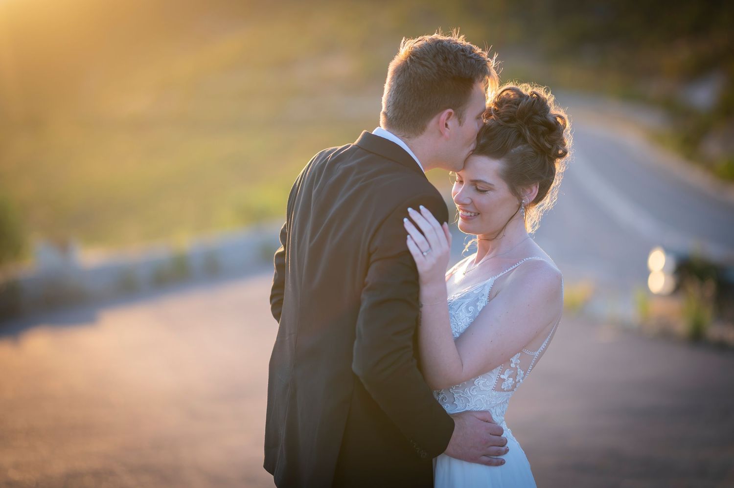 Two figures embrace tenderly while silhouetted against a blurred golden sunset background on a road.