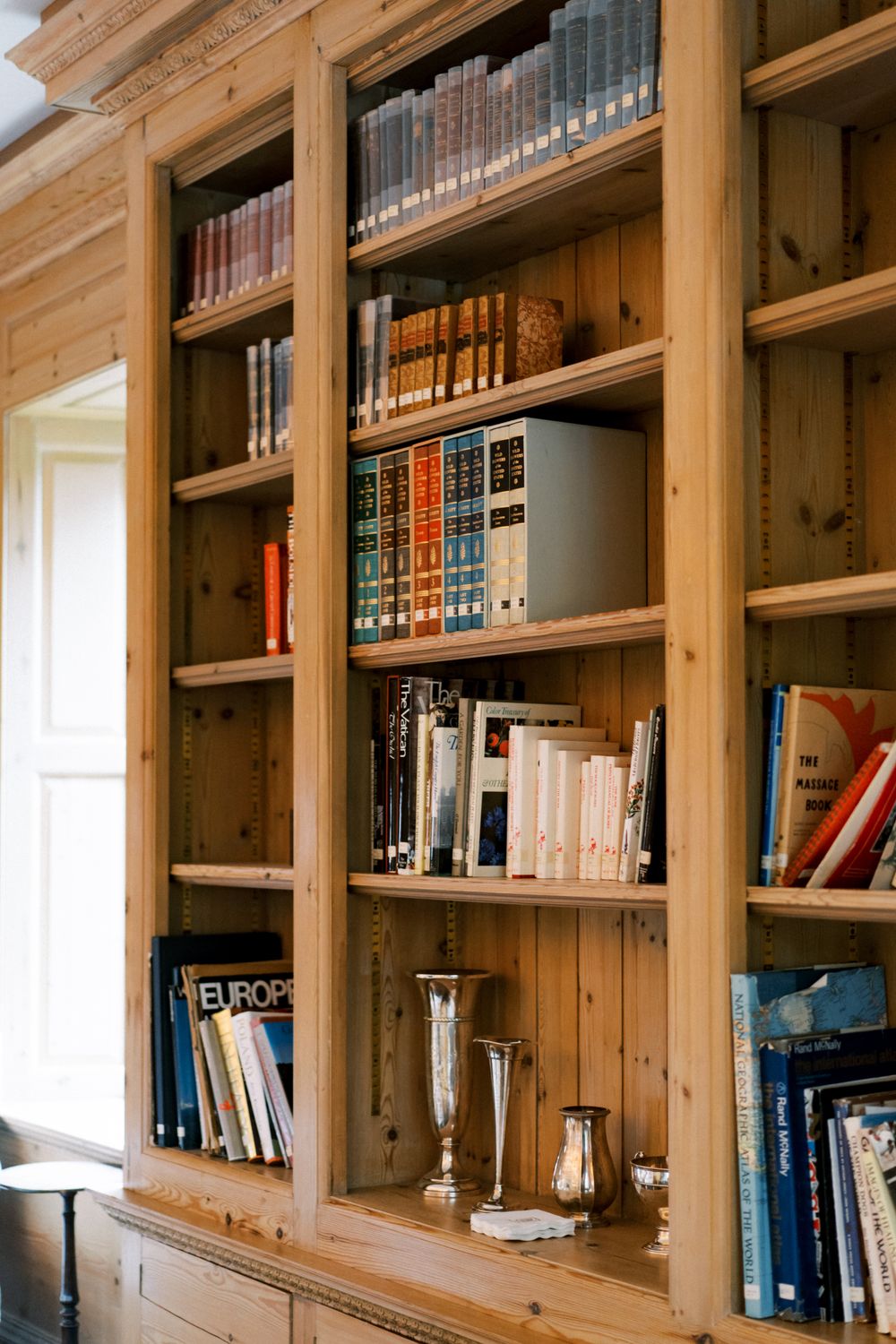 Built-in wooden library shelves filled with books line the walls of a rustic room with natural lighting.