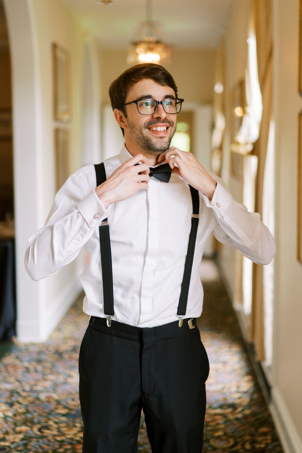 A joyful groom in suspenders and black pants adjusts his bow tie while getting ready in an elegant hotel hallway.