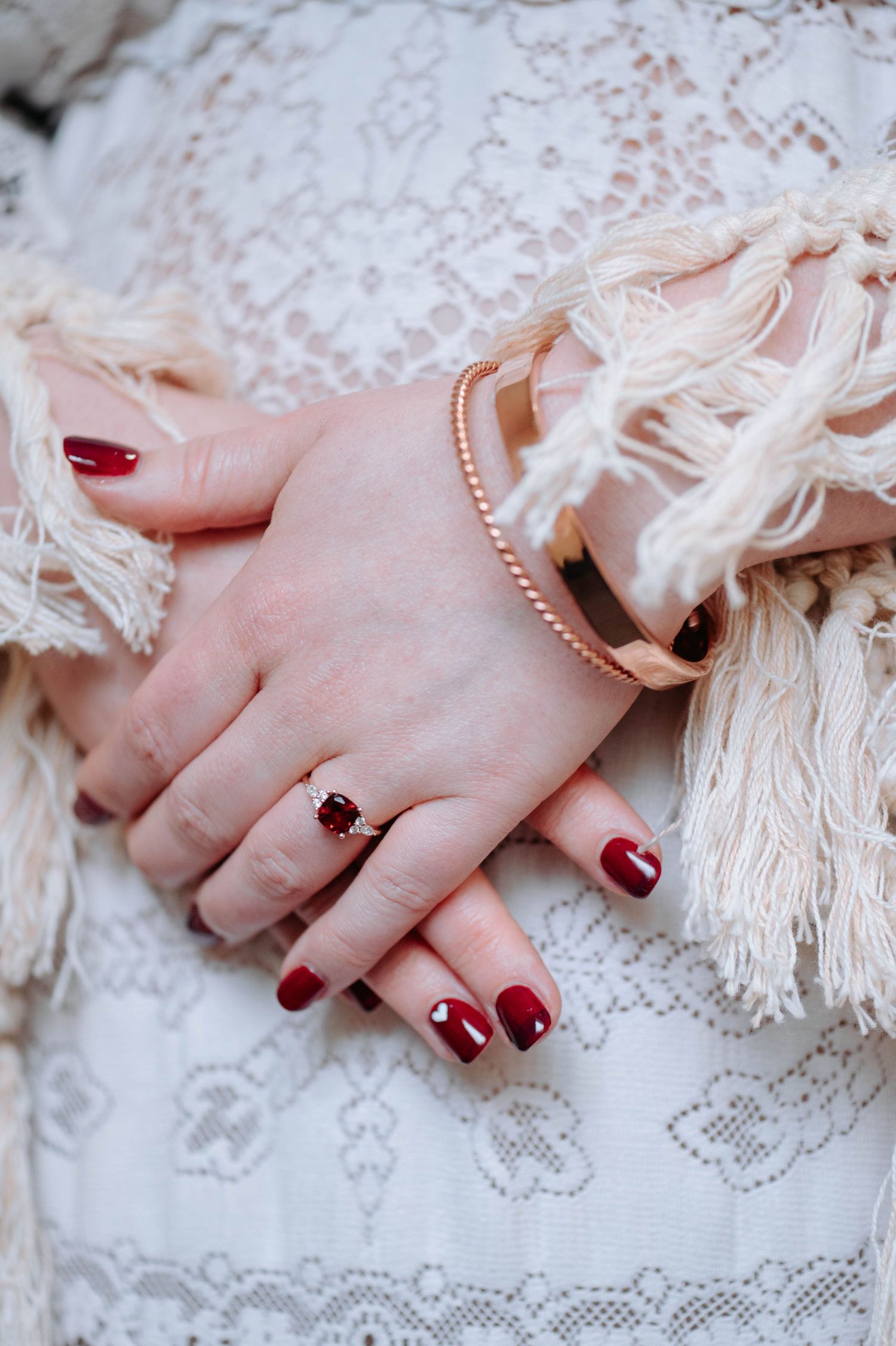 Close-up of a hand with red nail polish wearing a delicate pearl bracelet and ruby ring against white lace fabric.