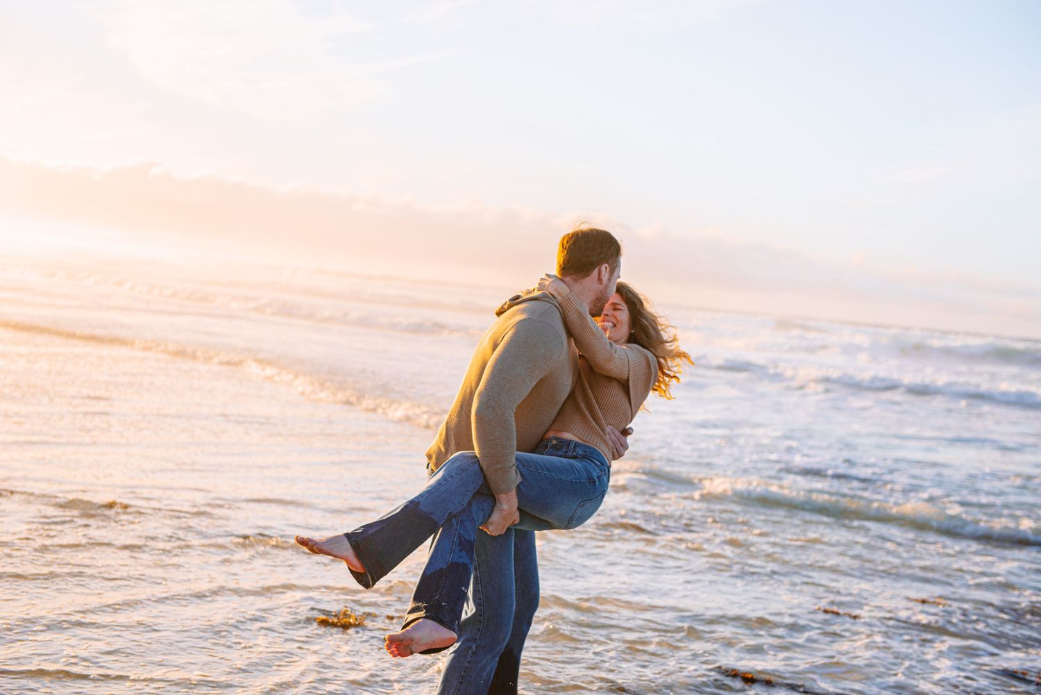 Two people embrace each other playfully on a sandy beach during sunset with ocean waves in the background.