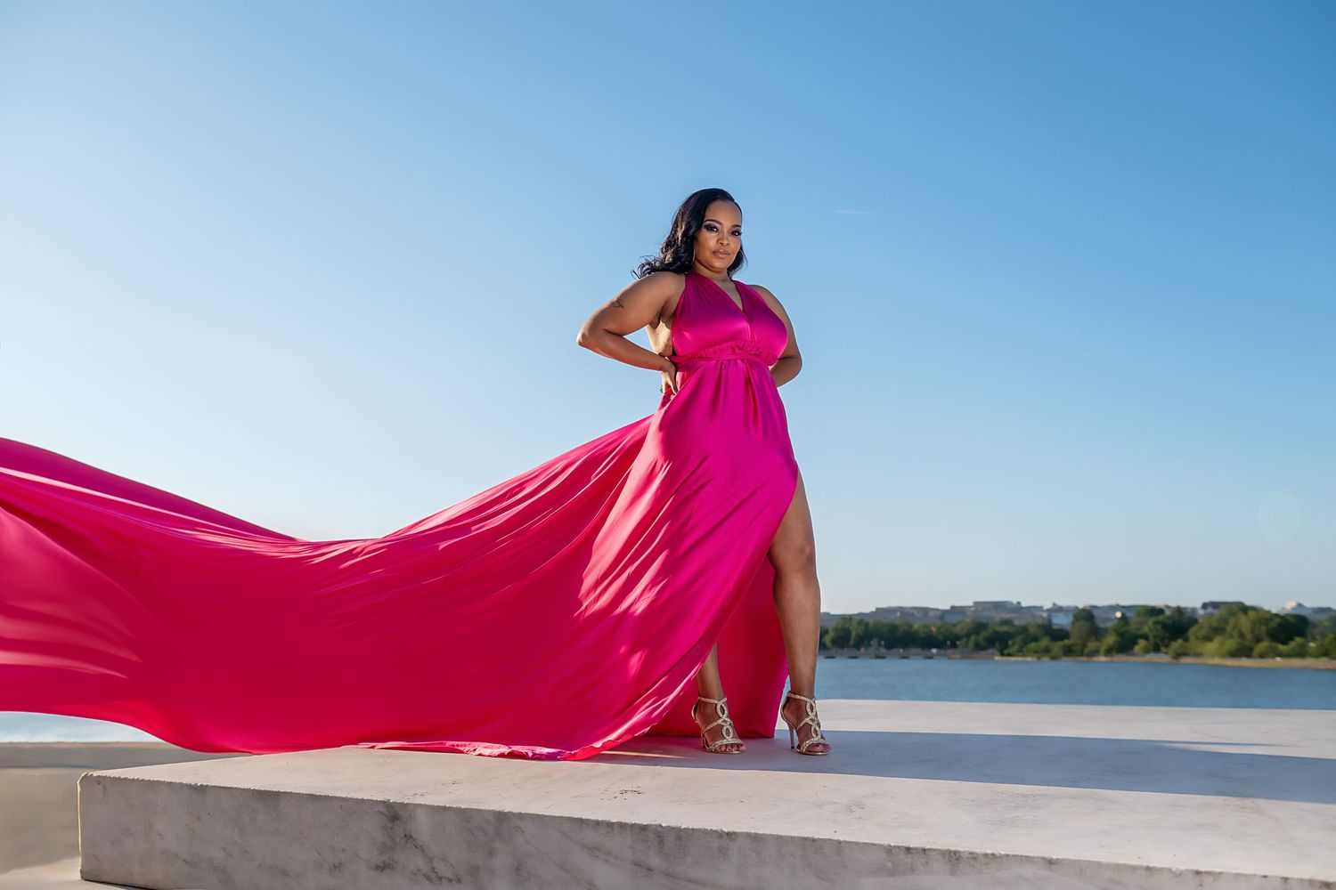 A person in a flowing hot pink dress poses gracefully along a beach shoreline on a bright sunny day.