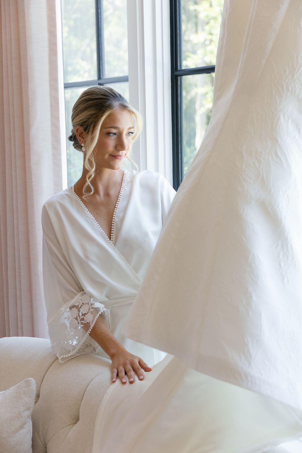 A person in a white robe sits by a window with soft natural light, creating an ethereal bridal preparation scene.