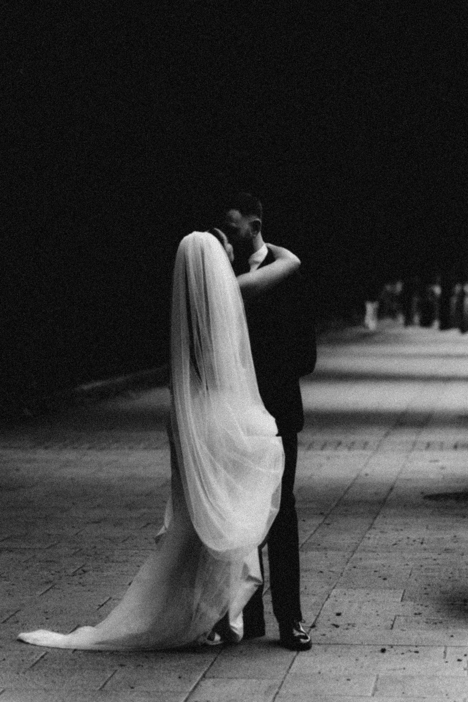 Black and white artistic wedding photograph showing bride's flowing veil and groom's shoes while walking on tiled floor.
