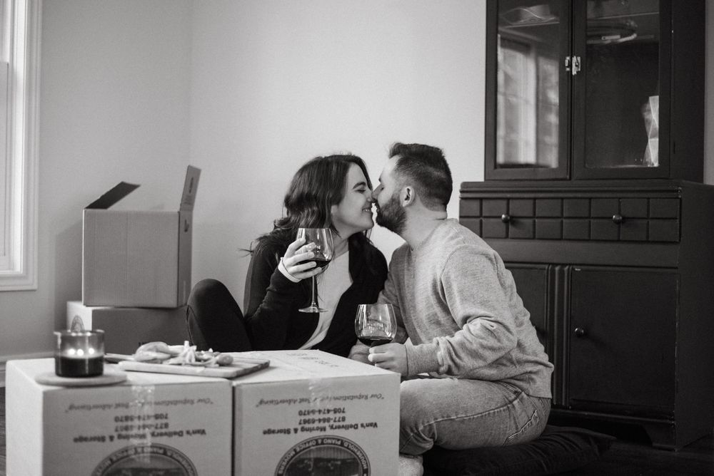 A couple shares an intimate moment while sitting among moving boxes in a black and white photograph.