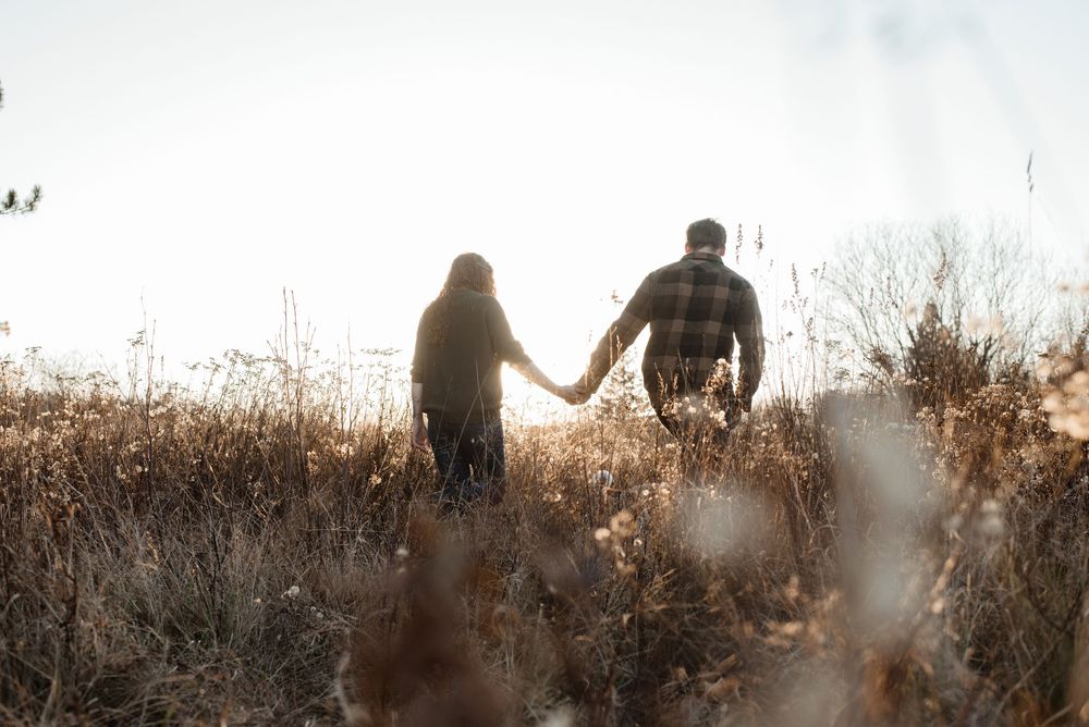 Silhouetted couple holding hands while walking through tall autumn grass at sunset.