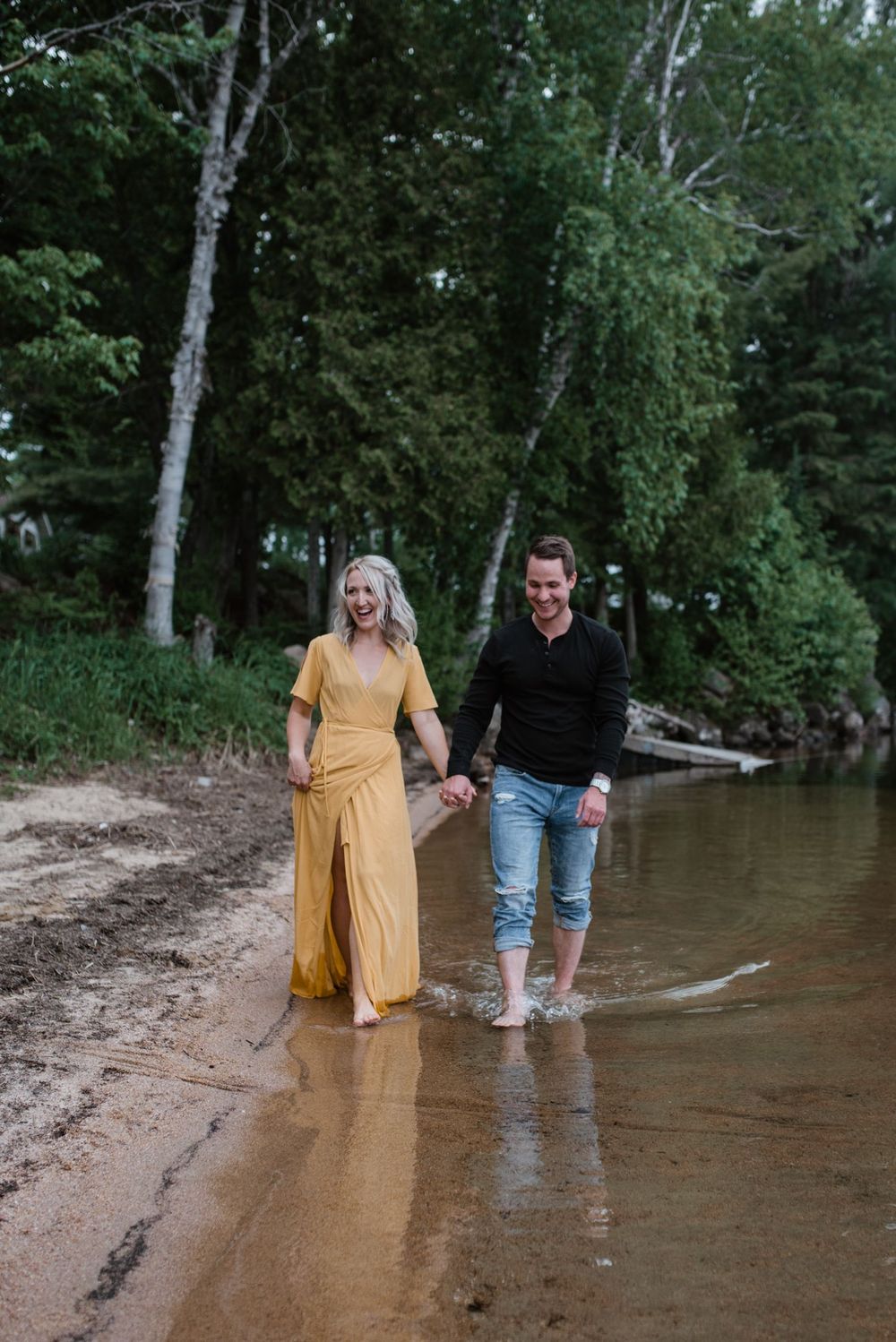 A couple walks together in the shallow water along a wooded shoreline on a summer day.