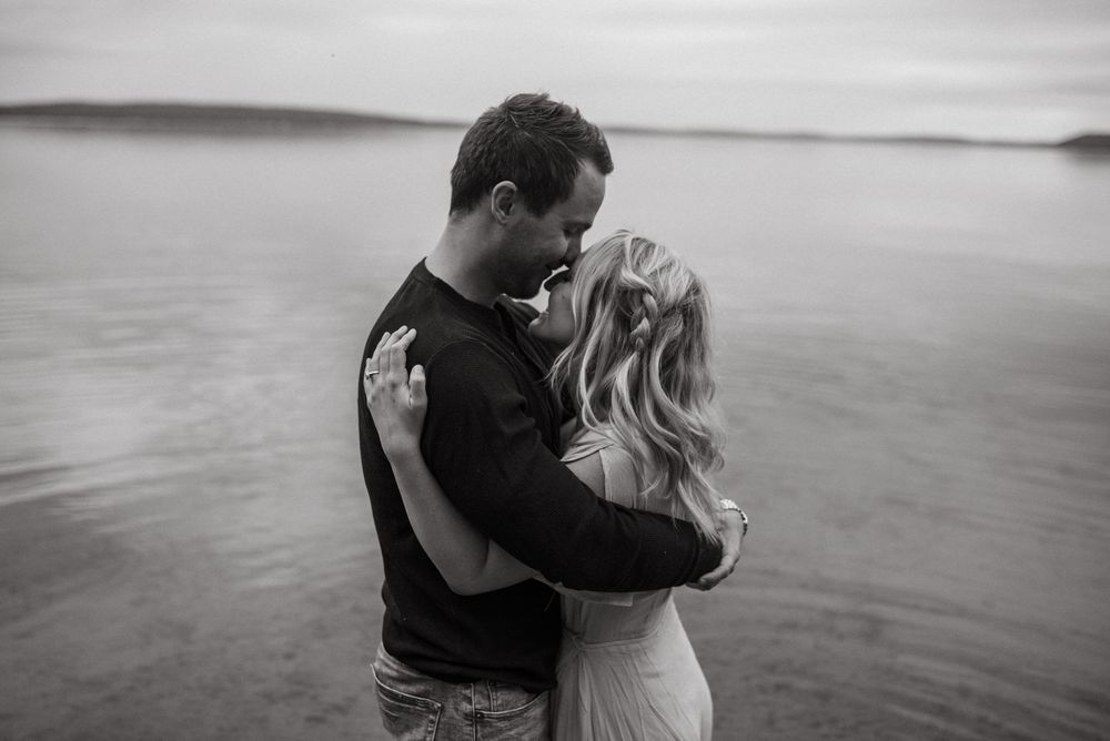 A romantic black and white portrait of a couple embracing by a serene waterfront at dusk.
