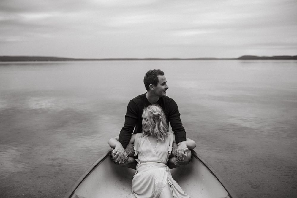 A couple sitting together in a canoe on a calm lake under an overcast sky in black and white.