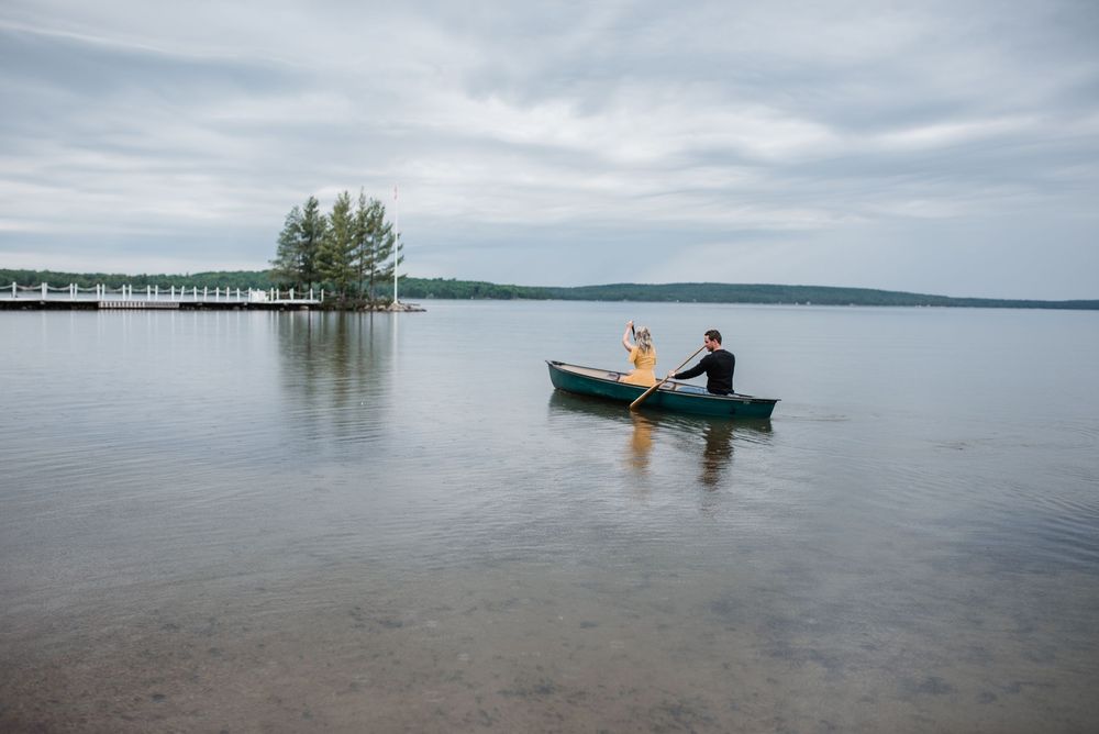 Two people paddle a canoe on a calm lake with a pier and trees visible in the background.
