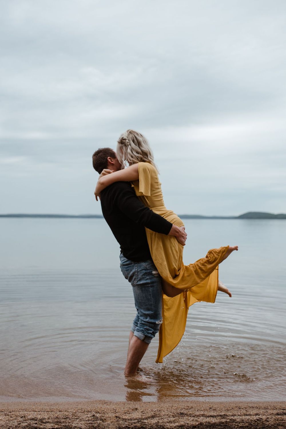 A couple embraces on a misty beach shore, with the woman wearing a flowing yellow dress as waves lap at their feet.