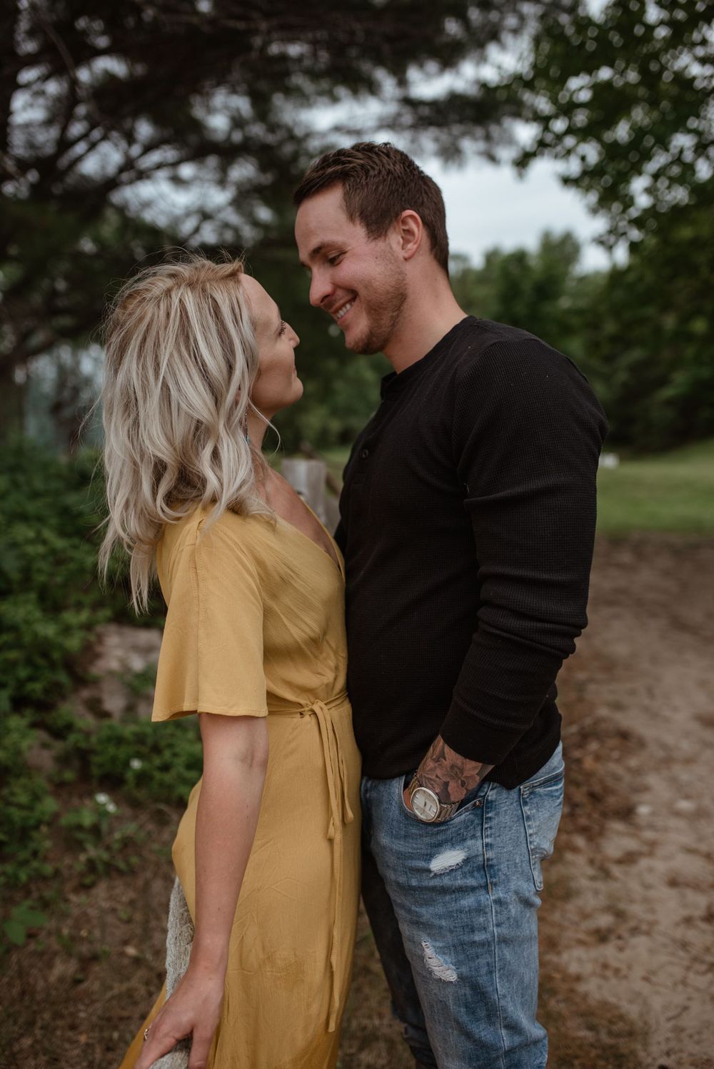 A couple shares an intimate moment on a dirt path surrounded by trees and greenery.