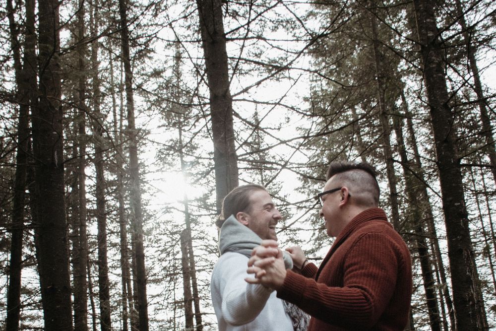 Two people share an intimate embrace while standing between tall evergreen trees in a misty forest setting.
