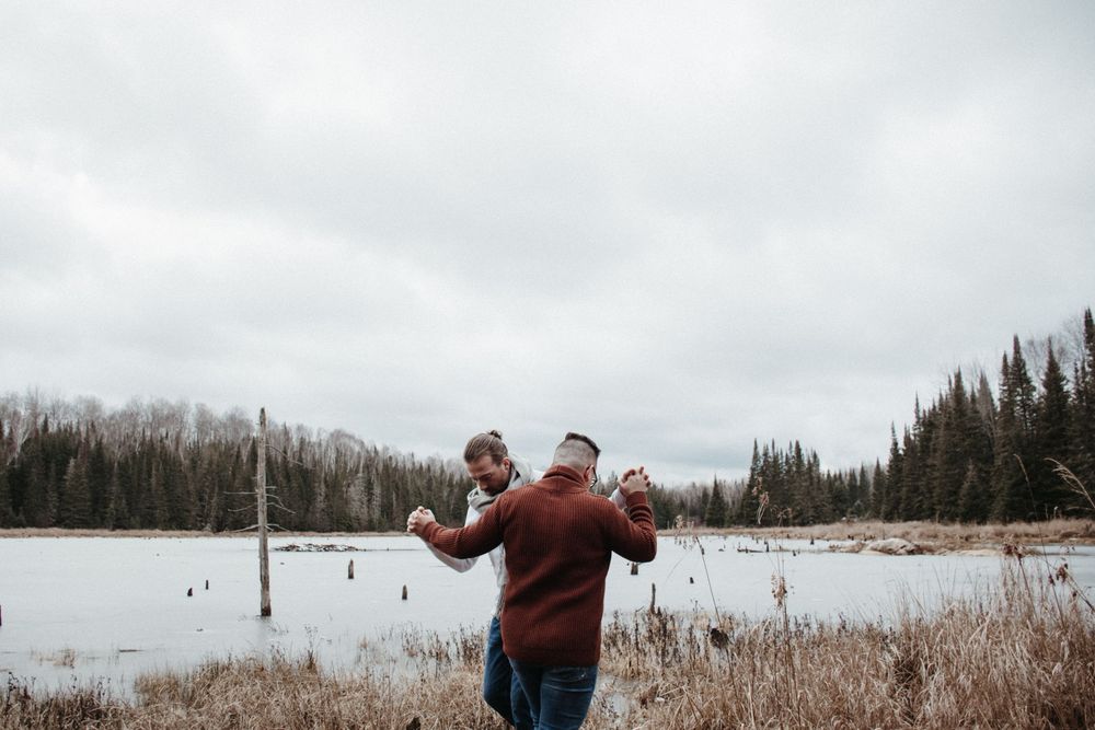 Person wearing a burgundy sweater stands at edge of a winter lake surrounded by evergreen trees and dried reeds.