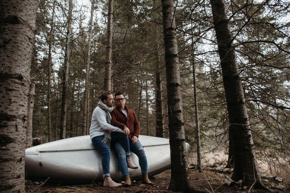 A couple sits together on a canoe in a moody forest setting surrounded by tall pine trees.