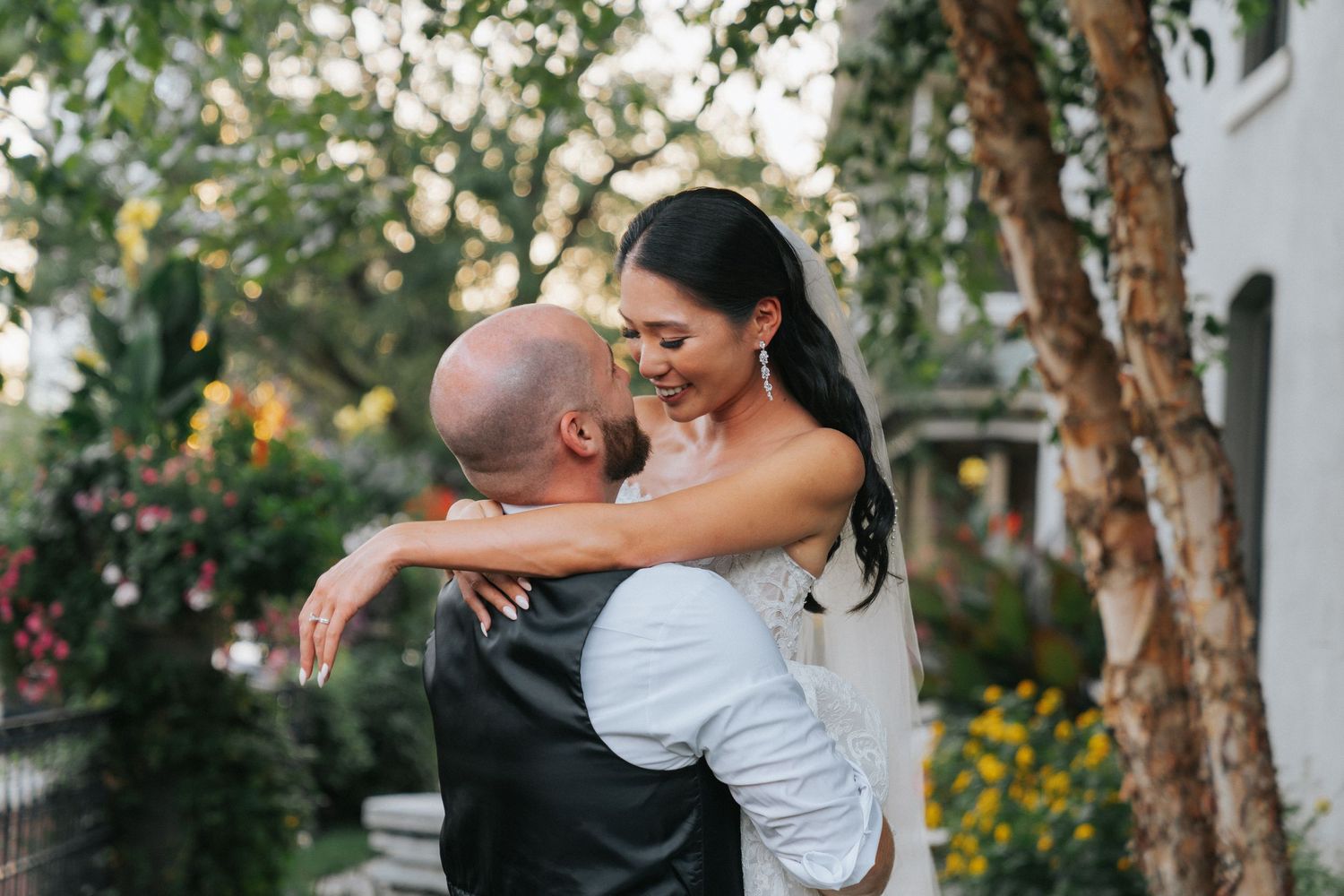 A couple shares a joyful embrace in a garden setting with trees and flowers in the background.