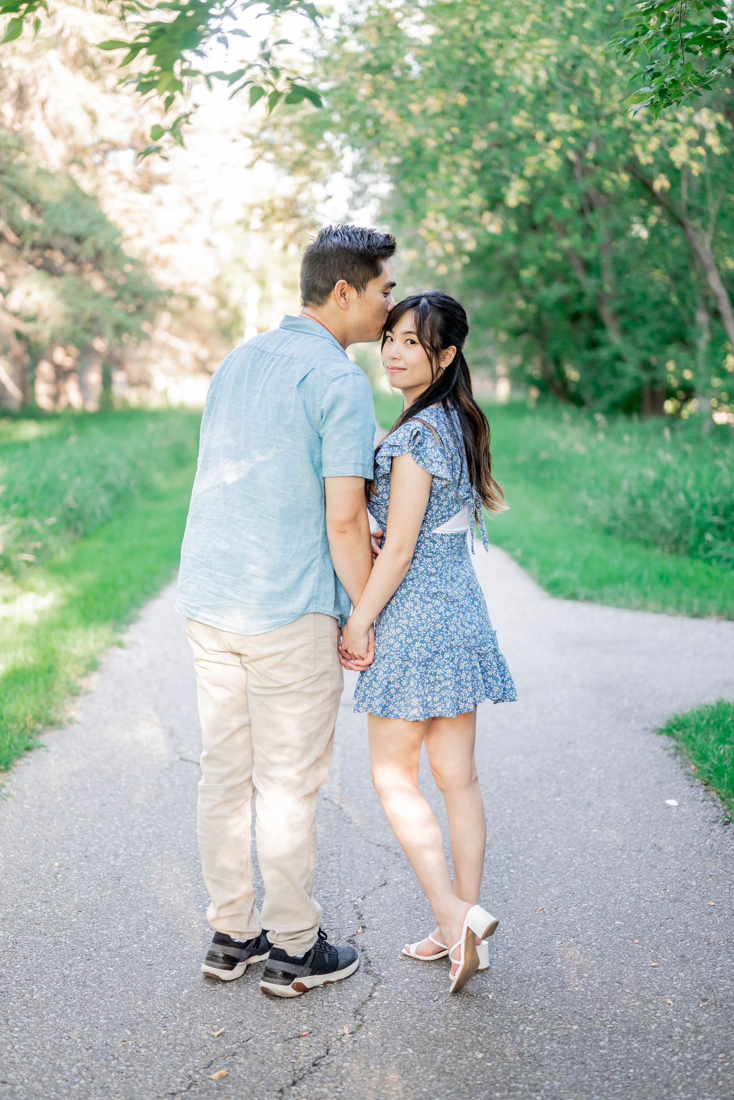 A couple in casual attire holding hands while walking on a sun-dappled path surrounded by lush greenery.