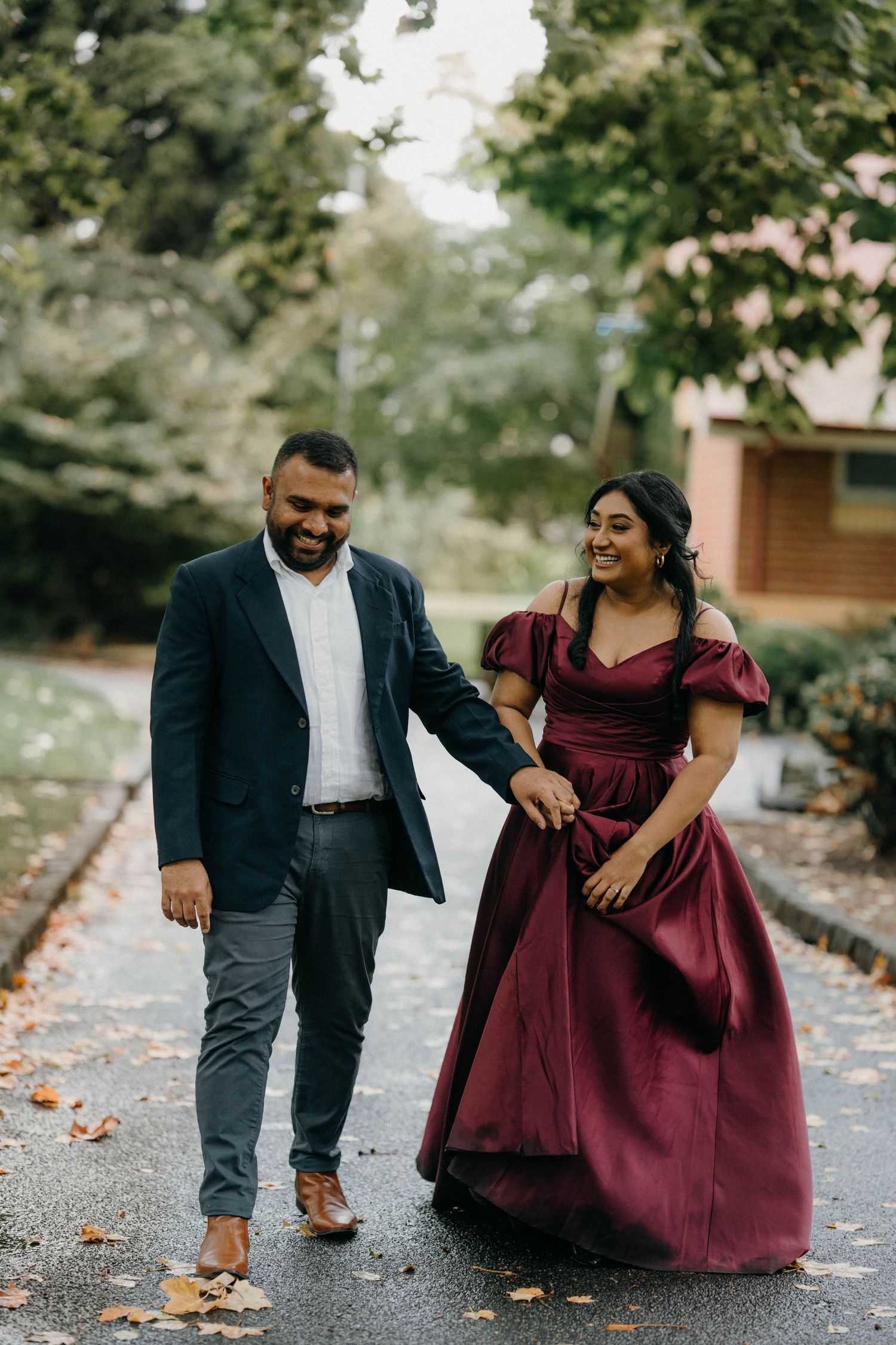 Couple walking and laughing together on a leaf-covered path, wearing formal burgundy dress and dark suit.