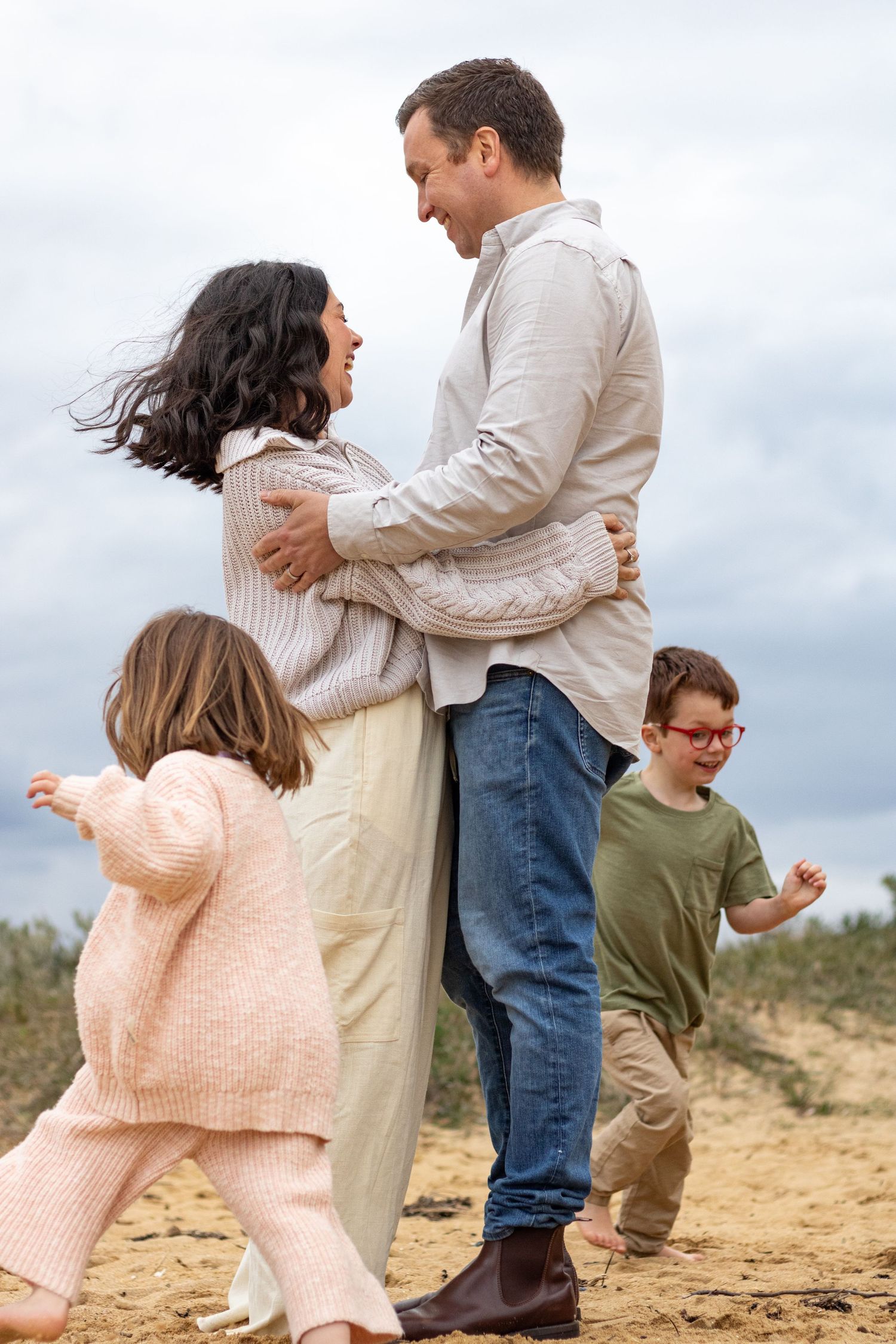 Family enjoying playful moments together on a sandy beach during an overcast day.