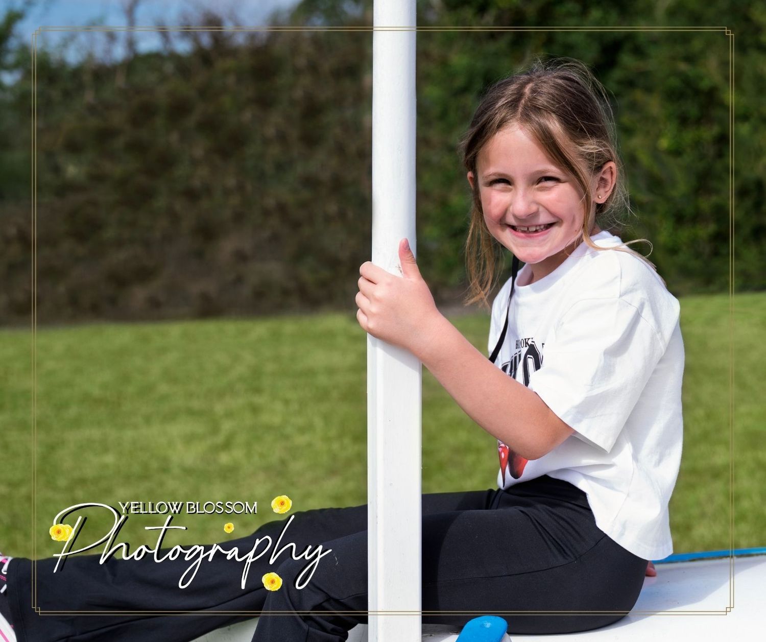 A youth athlete in a white jersey sits by a soccer goal post on a sunny day at a sports field.