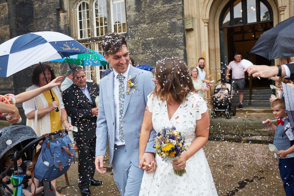Wedding couple exits stone church while guests celebrate with confetti and umbrellas.