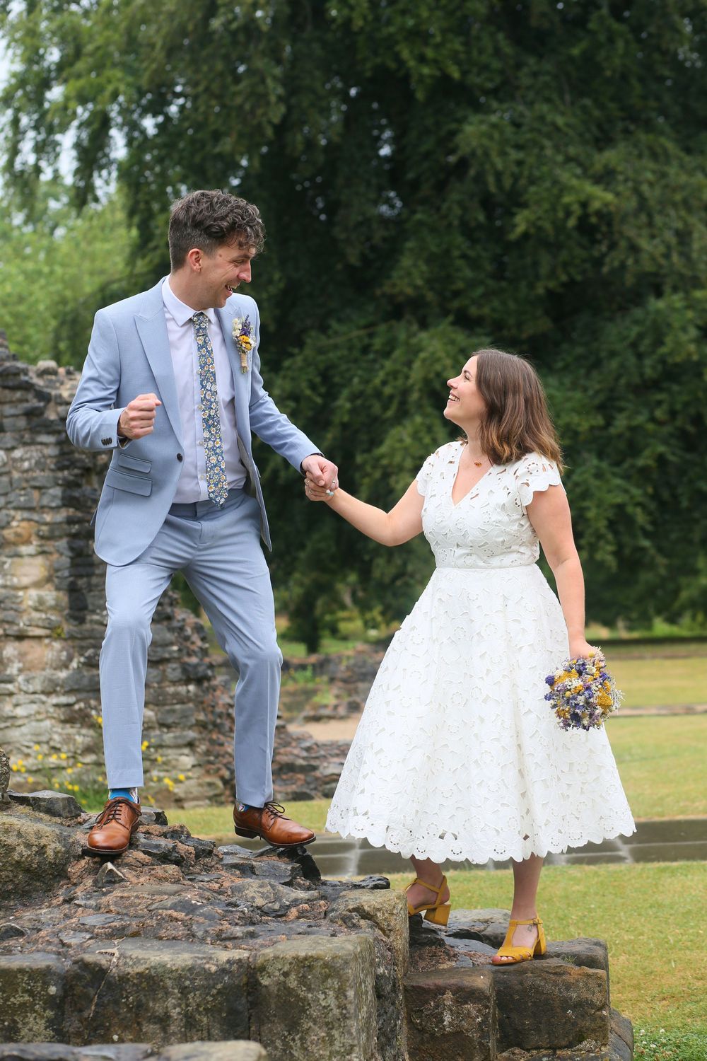 Playful wedding couple poses on stone steps in light blue suit and tea-length white dress.