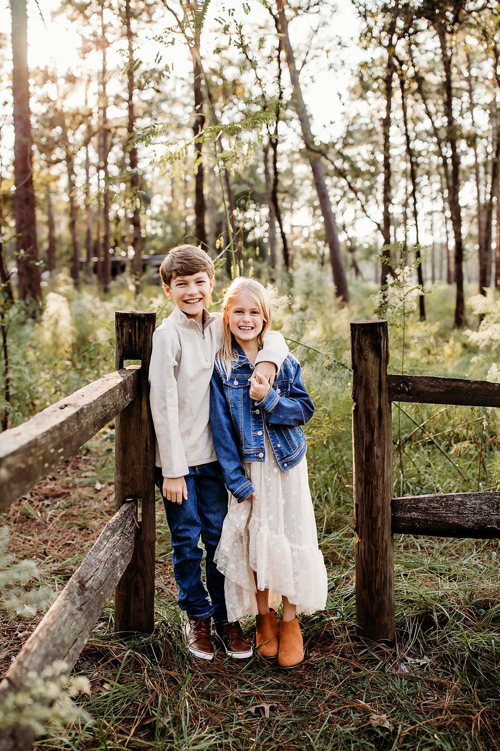 Two children share a sweet moment while leaning against a rustic wooden fence in a sunlit pine forest.