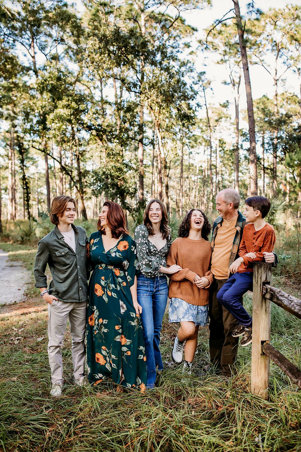 A group of friends share laughter while leaning against a wooden fence in a sun-dappled forest setting wearing autumn attire.