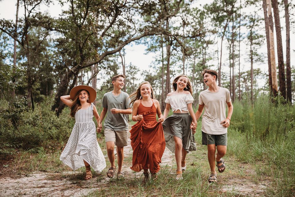 A group of young people laugh and run together on a sandy forest path during a bright summer afternoon.