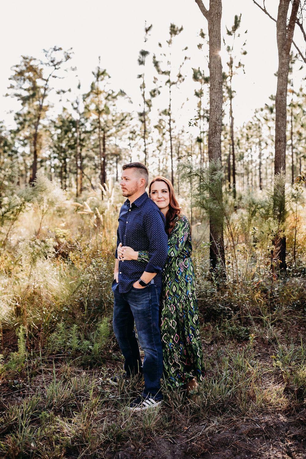 A couple embraces while standing among tall pine trees and natural foliage in a forest setting.