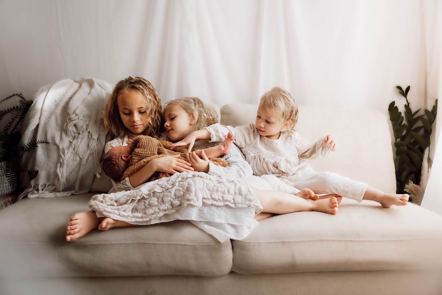 Three siblings relaxing together on a white couch with soft natural lighting, holding their newborn baby brother.