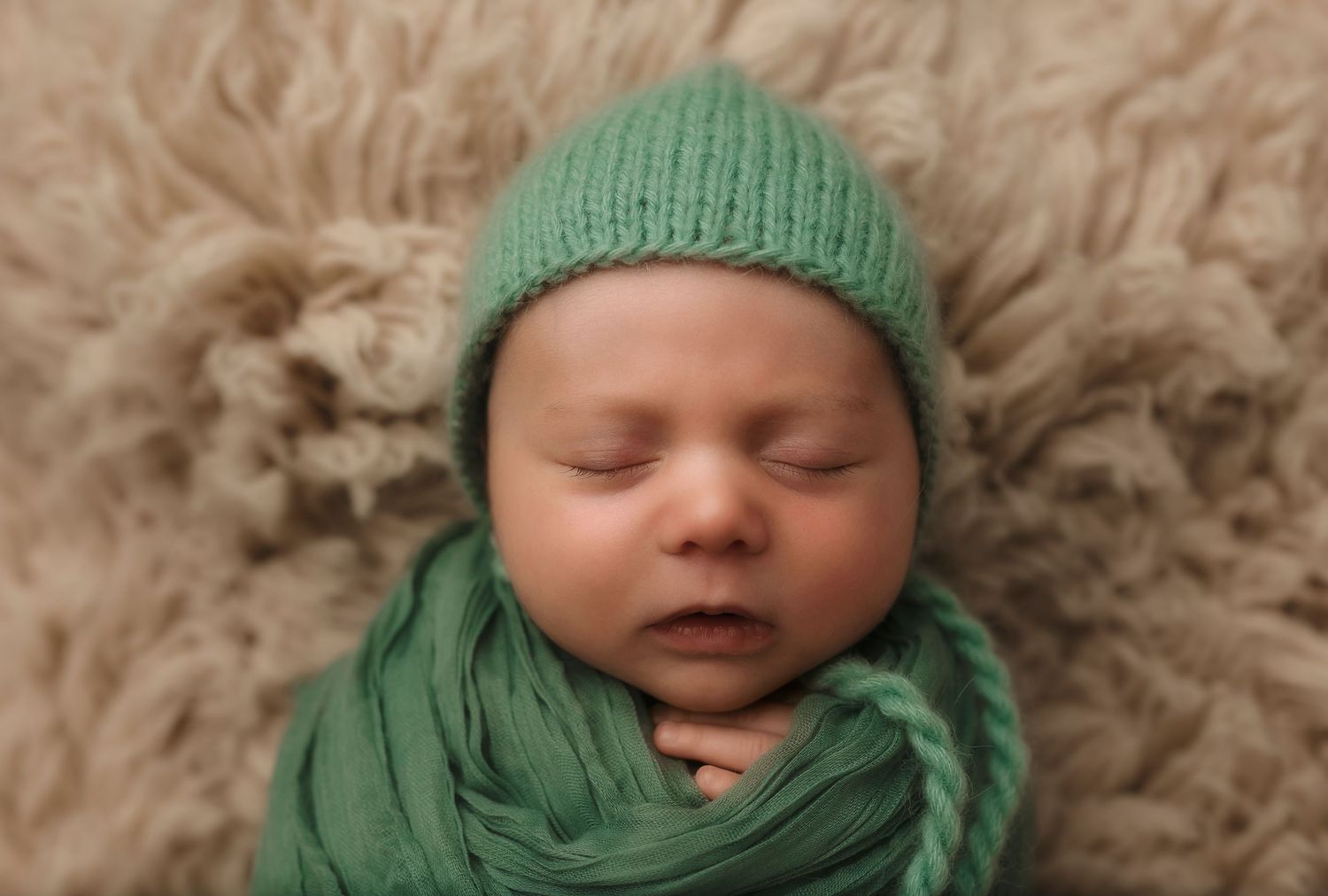 Newborn boy sleeps soundly in minty green wrap and matching bonnet on a wool rug, captured by Charlottesville baby photographer.