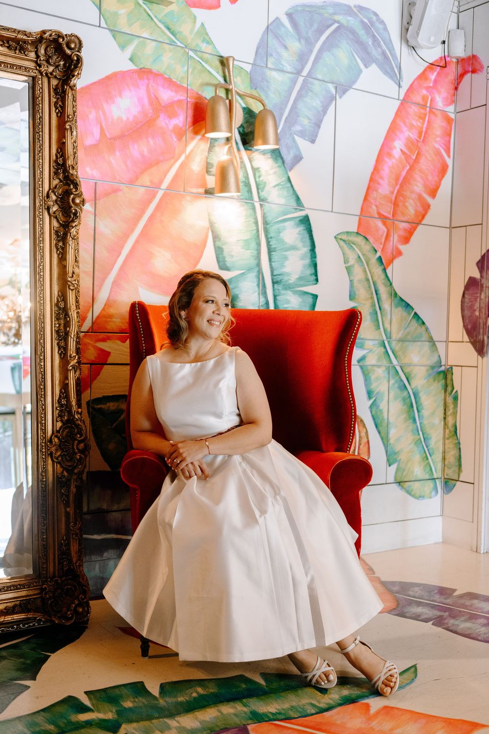 A white wedding dress against tropical leaf decor and red velvet seating in an elegant interior setting.