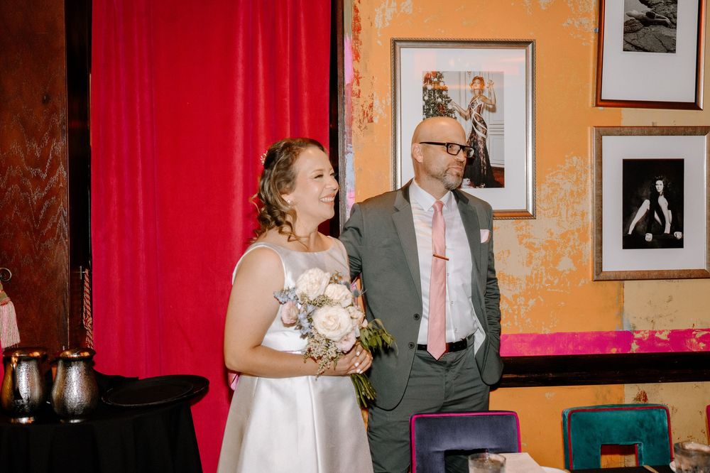 A wedding couple and guests socialize in an orange-walled room decorated with framed artwork and red curtains.