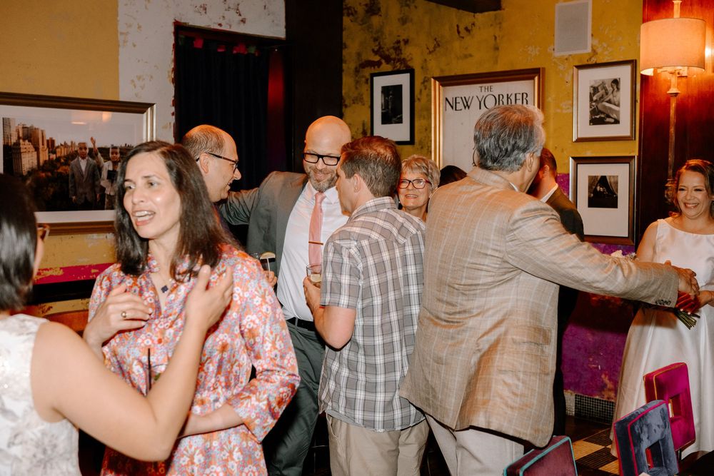Wedding guests mingle and dance at a reception in a room with framed artwork and warm lighting.