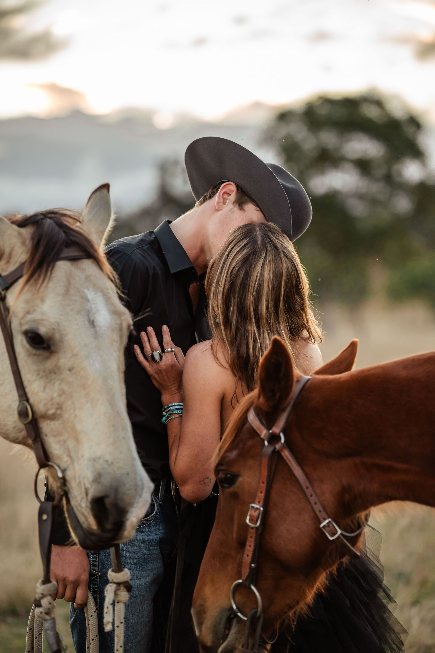 A romantic couple sharing a moment between two horses at sunset in a rustic countryside setting.