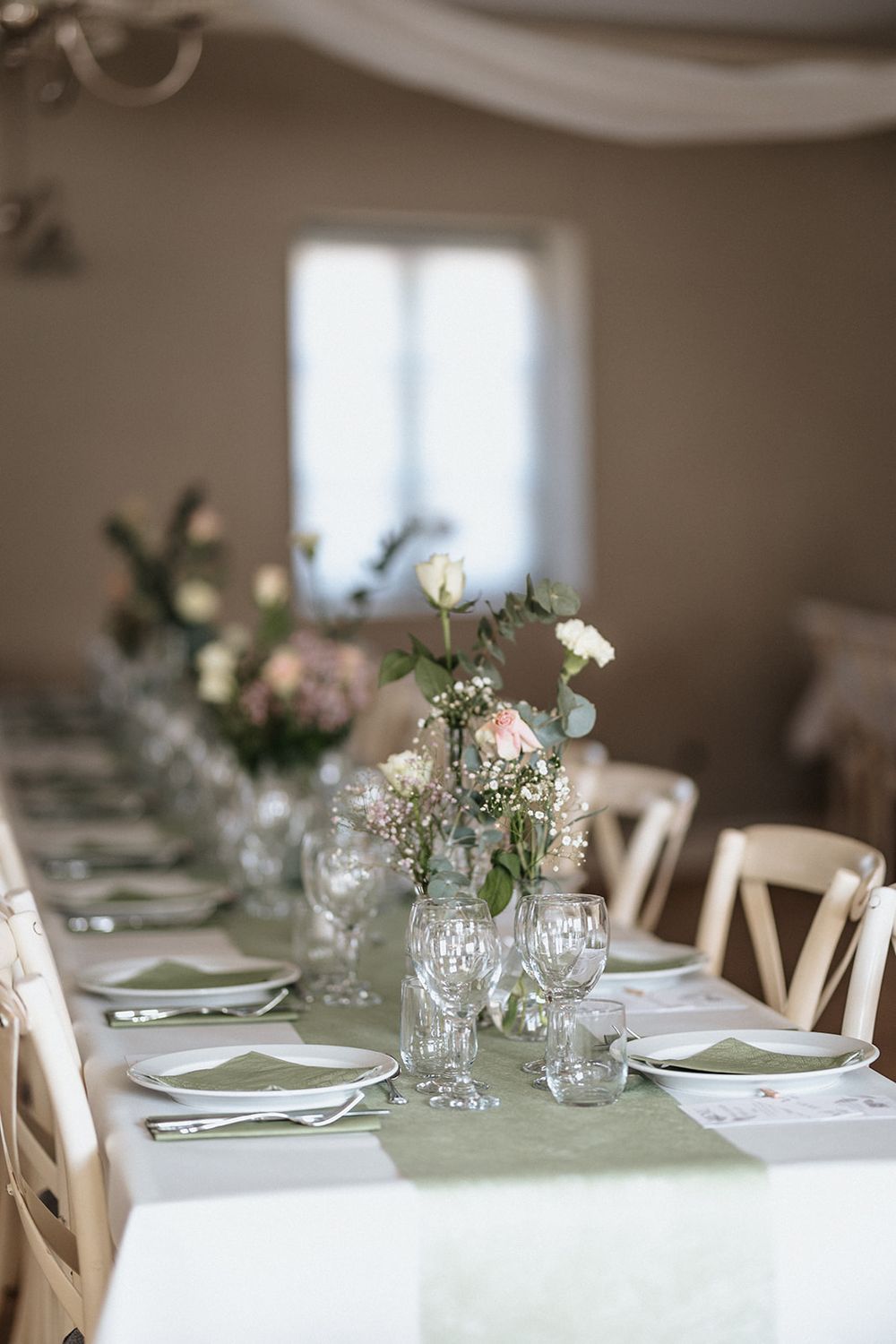Wedding reception table decorated with white floral centerpieces and sage green linens in a bright room.