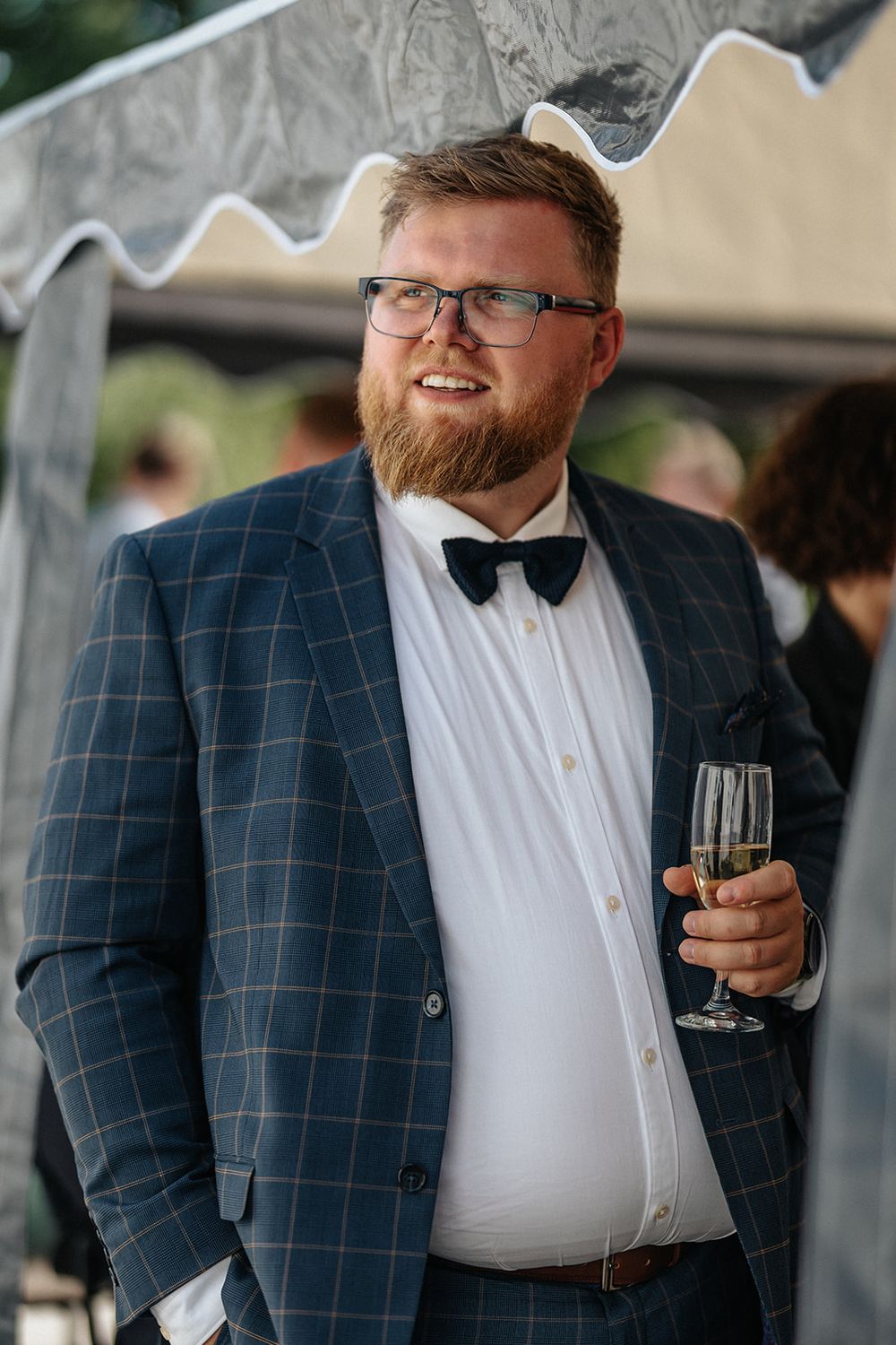 Wedding guest in a checkered navy blazer and bow tie holds a drink while enjoying outdoor reception festivities.