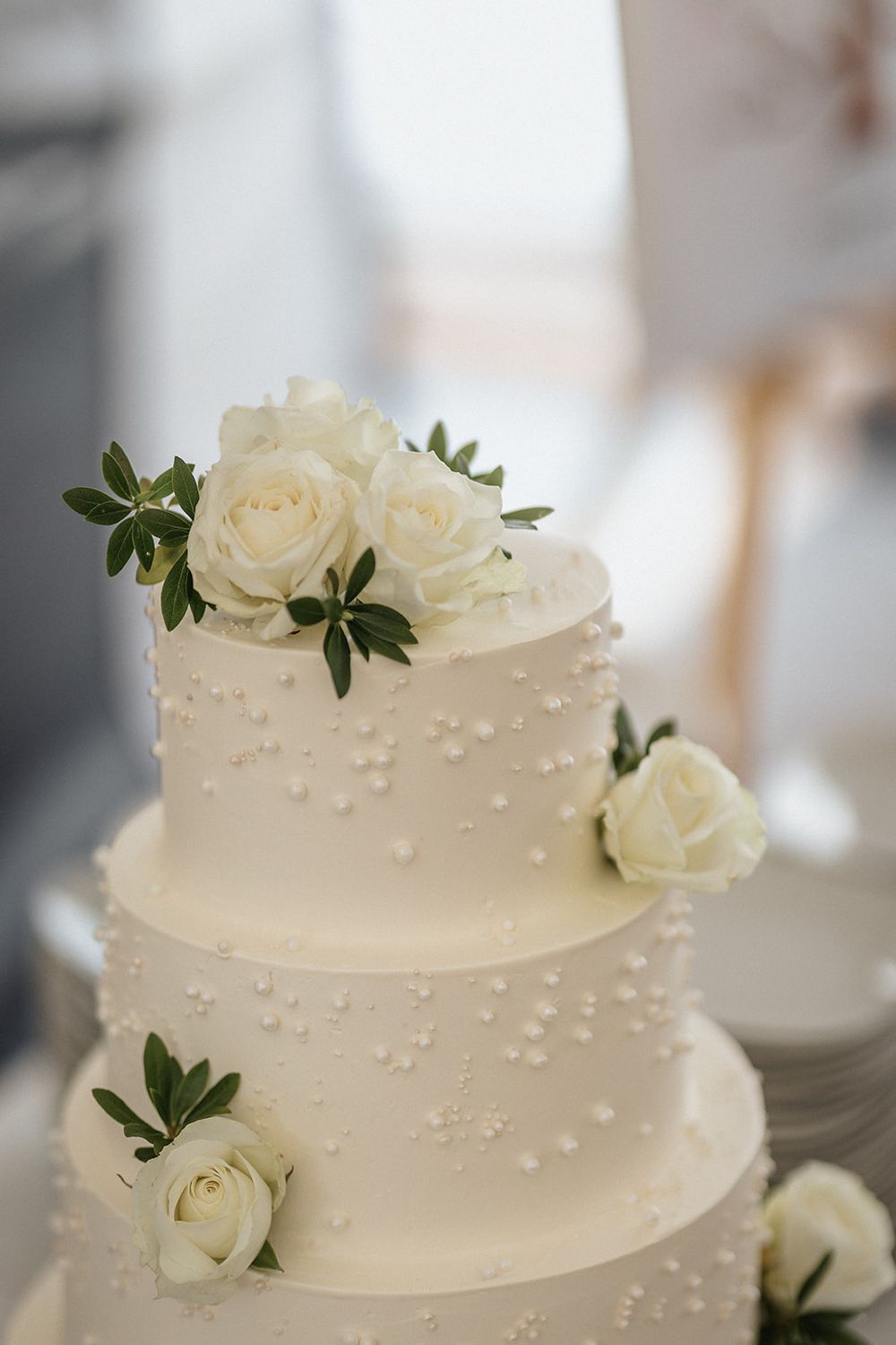 Elegant white wedding cake decorated with delicate pearl accents and fresh white roses on each tier.