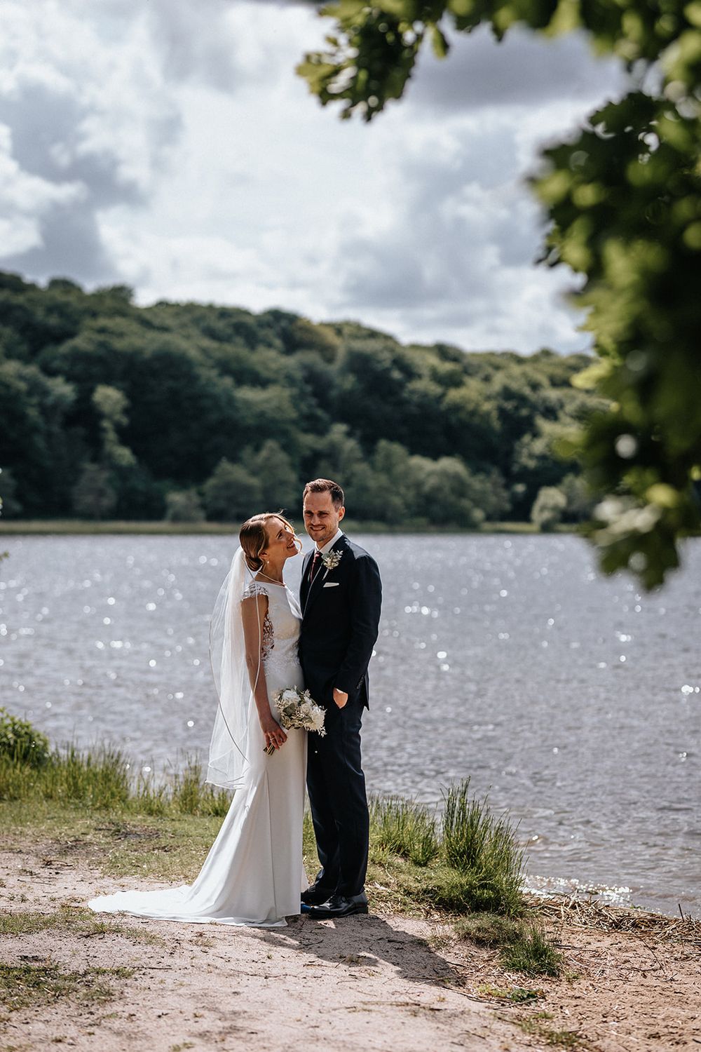 A bride and groom share a romantic moment by a scenic lake shore with lush hills and sparkling water in the background.