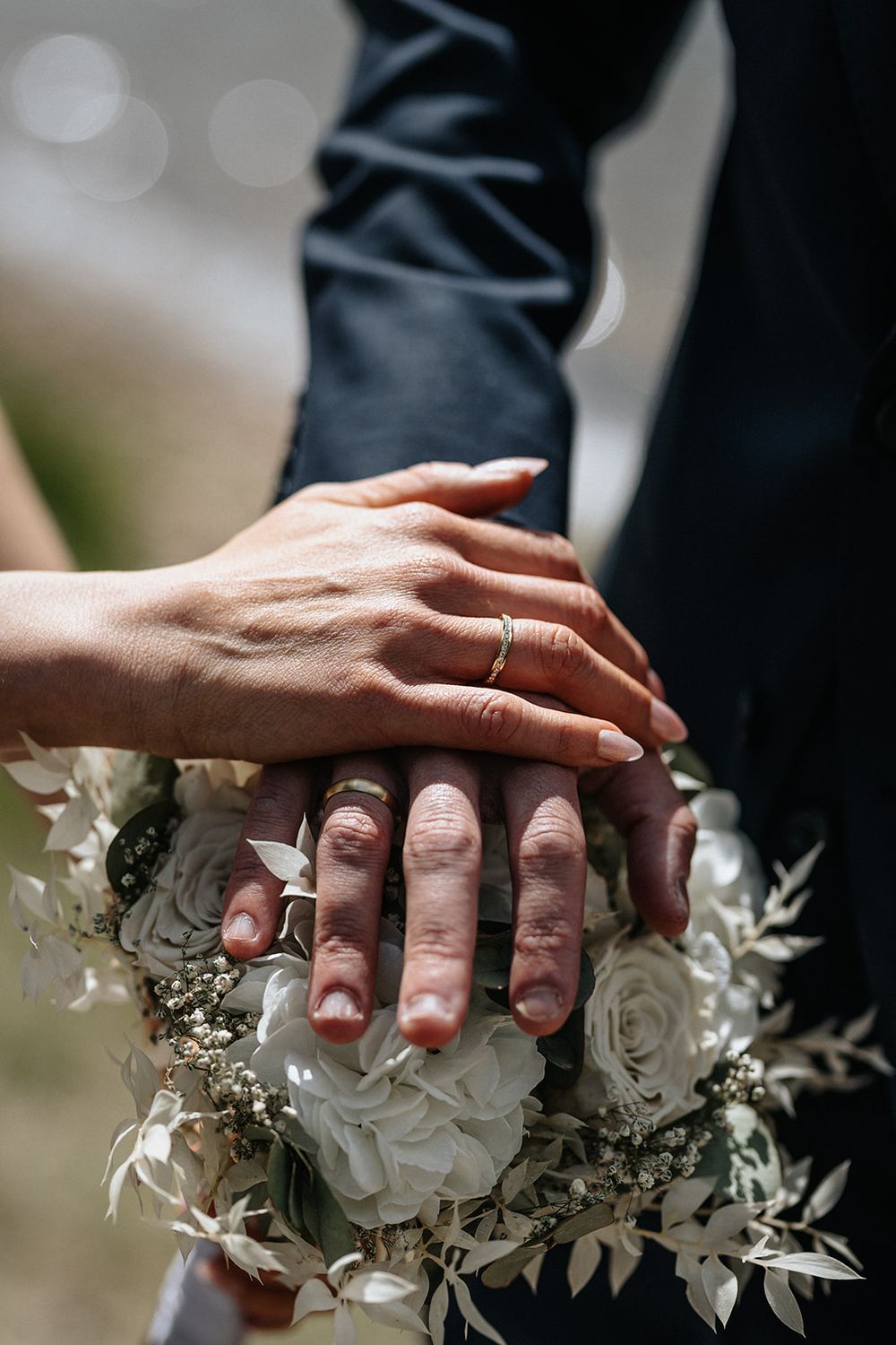 Newlywed couple's hands with wedding rings rest on a white rose and greenery bouquet in an intimate moment.
