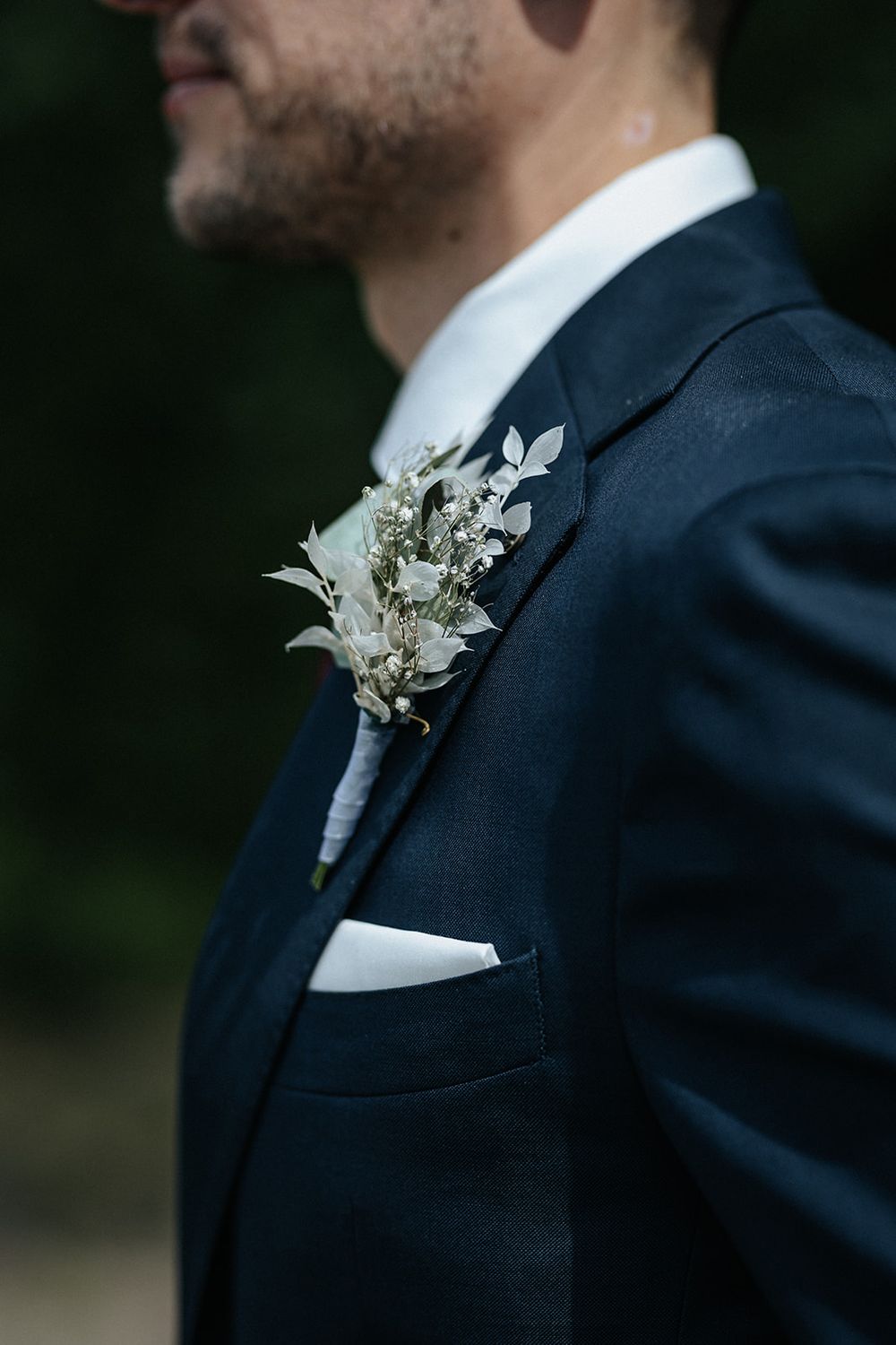 Close-up detail of a white and green floral boutonniere pinned to a navy blue suit jacket with white pocket square.