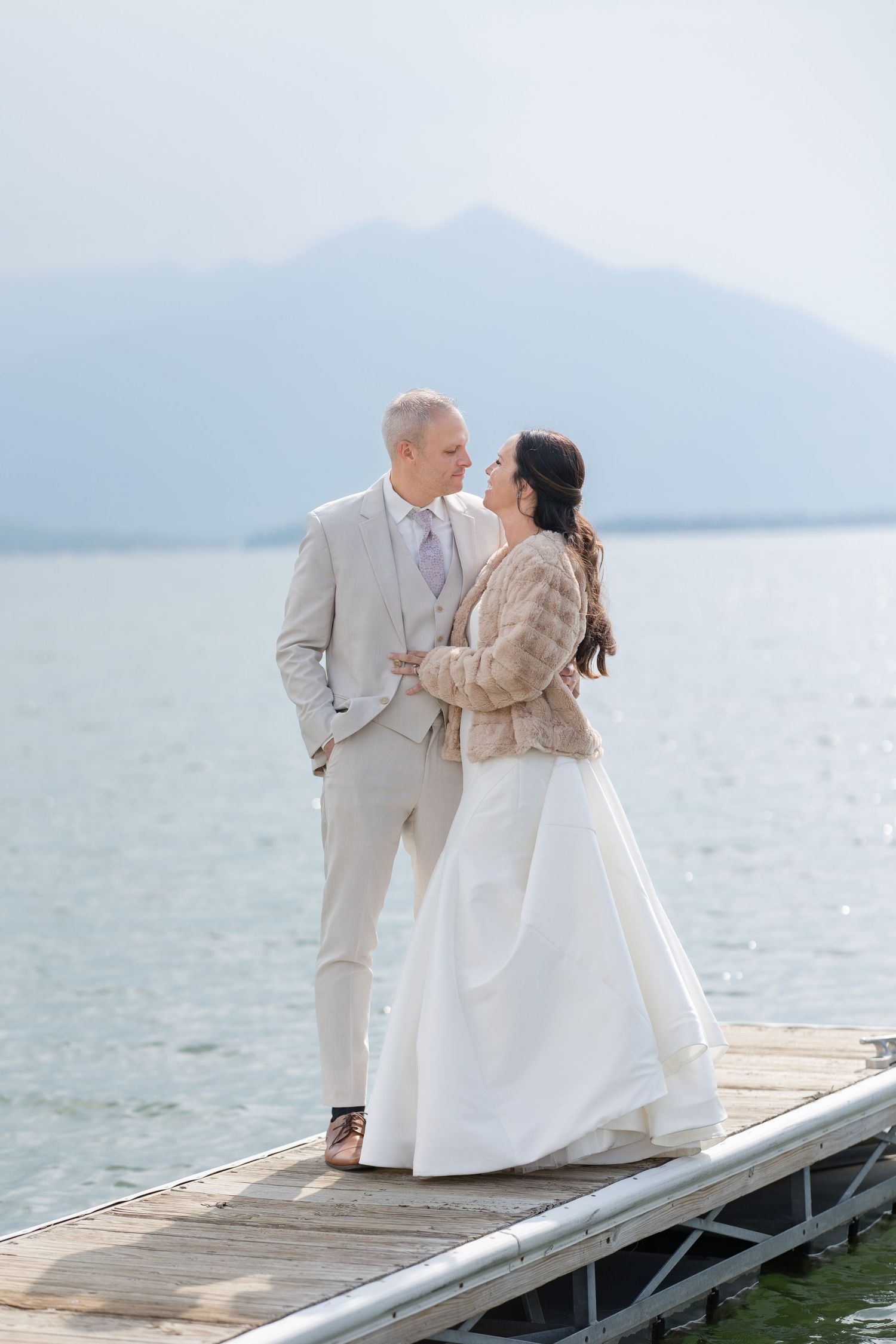 A couple in wedding attire stands on a dock overlooking misty mountains and lake on their special day.