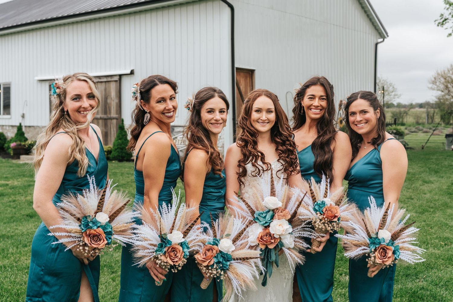 Bridesmaids in teal dresses pose together outdoors near a rustic barn.