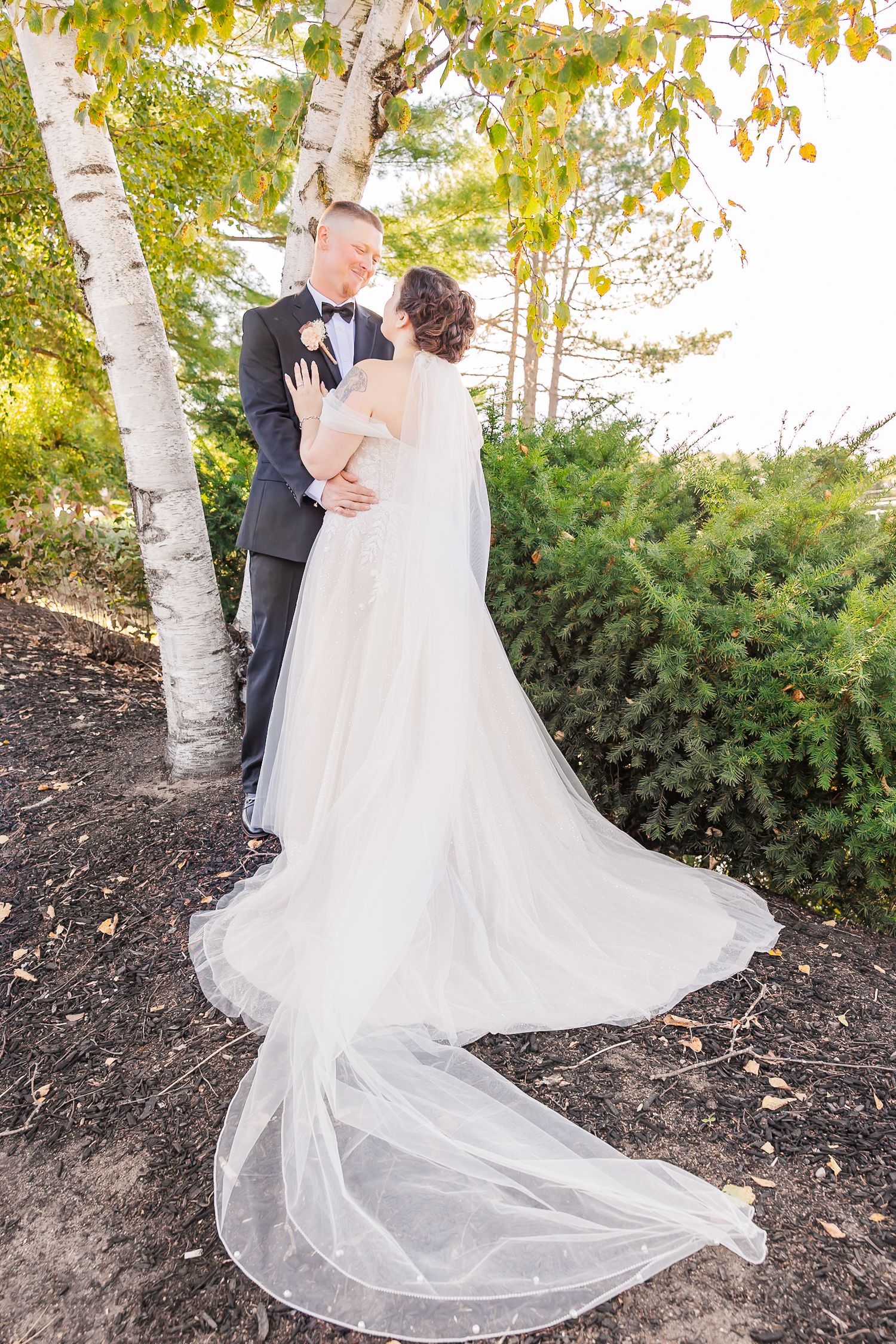 A flowing white wedding dress train trails beautifully across autumn leaves beneath birch trees in an outdoor setting.